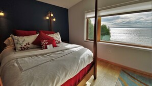 A bedroom with a bed, a nightstand, a lamp, a window with a view of the water, and a wooden floor. - LaBarbe Bleu on Lake Michigan (Saint Ignace)