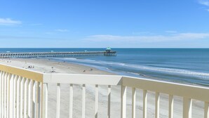 A beach with a pier extending into the water, a clear sky, and a white balcony railing. - Stay @ Paradise Peak on Folly Beach (Folly Beach)