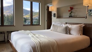 Un grand lit avec du linge de lit blanc et un plaid beige, deux lampes de chevet, une tête de lit en bois, une petite table avec une chaise, et une vue sur un paysage montagneux par de grandes fenêtres.