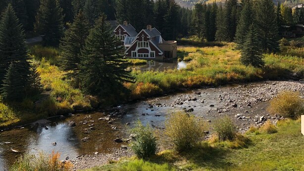 Une maison située en bord de rivière, entourée d’arbres, avec une montagne en toile de fond.