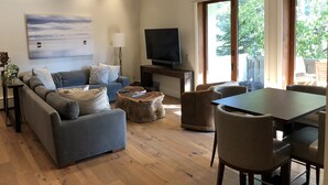 Un salon moderne avec un canapé gris, une table basse et une table à manger avec des chaises. Un téléviseur à écran plat est fixé au mur, ainsi qu’une lampe et une grande fenêtre donnant sur un paysage verdoyant.