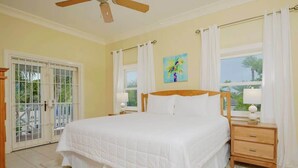 Une chambre à coucher comprenant un lit, une table de chevet, une fenêtre avec des rideaux, un ventilateur de plafond et une porte donnant sur un espace extérieur.