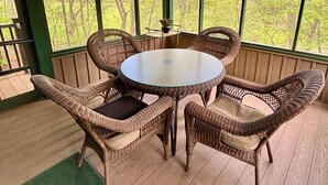 Un espace extérieur aménagé, abrité et doté de chaises en osier et d’une table en verre, entouré de grandes fenêtres offrant une vue sur les arbres.