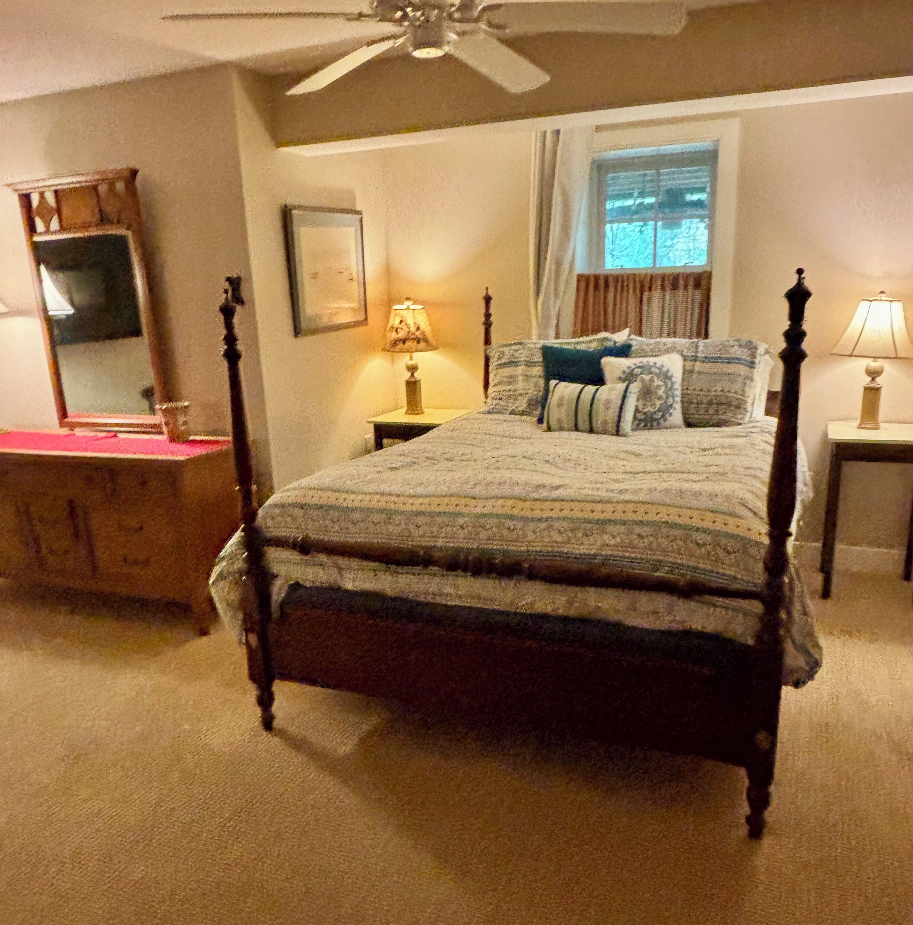 A bedroom with a four-poster bed, a nightstand with a lamp, a dresser, and a ceiling fan.