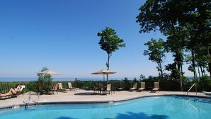 A swimming pool with lounge chairs and umbrellas, surrounded by trees and a clear view of the ocean. - Magnificent Water View Condo (Egg Harbor)