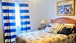 A bedroom with a bed, curtains, a ceiling fan, a nightstand, a lamp, and a painting on the wall. - Located in desirable Ocean Walk, short walk to ocean and village (St Simons Island)