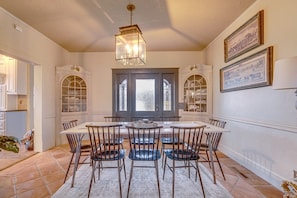 Uma sala de jantar com uma mesa grande, cadeiras e um candelabro. Há duas janelas com cortinas e uma porta com painéis de vidro.