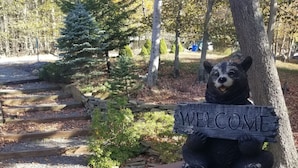 A wooden bench with a bear sculpture holding a 'WELCOME' sign, surrounded by trees and a gravel path.