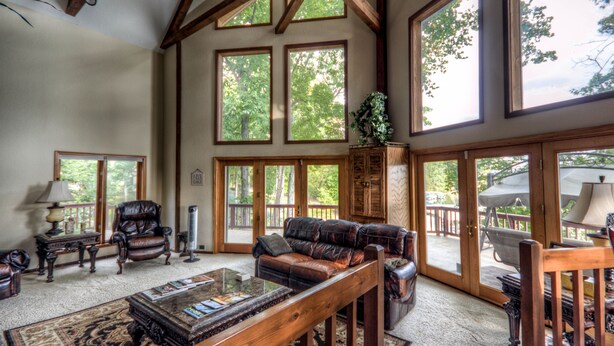A spacious living room with a large leather sofa, a coffee table, and a fireplace. There are multiple windows providing natural light and views of the outdoors.