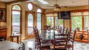 A dining area with a large table, wooden chairs, and a television. There are large windows with views of a lake.