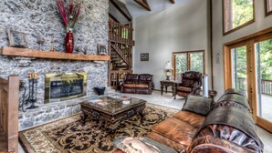 A spacious living room with a stone fireplace, leather furniture, and large windows offering a view of the outdoors.