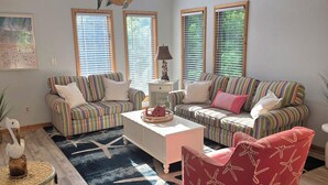 A living room with striped sofas, a white coffee table, a red chair, and a ceiling fan. - Private Whalehead Beach Retreat - Pool, Hot Tub & 5-Minute Walk to the Ocean! (Corolla)