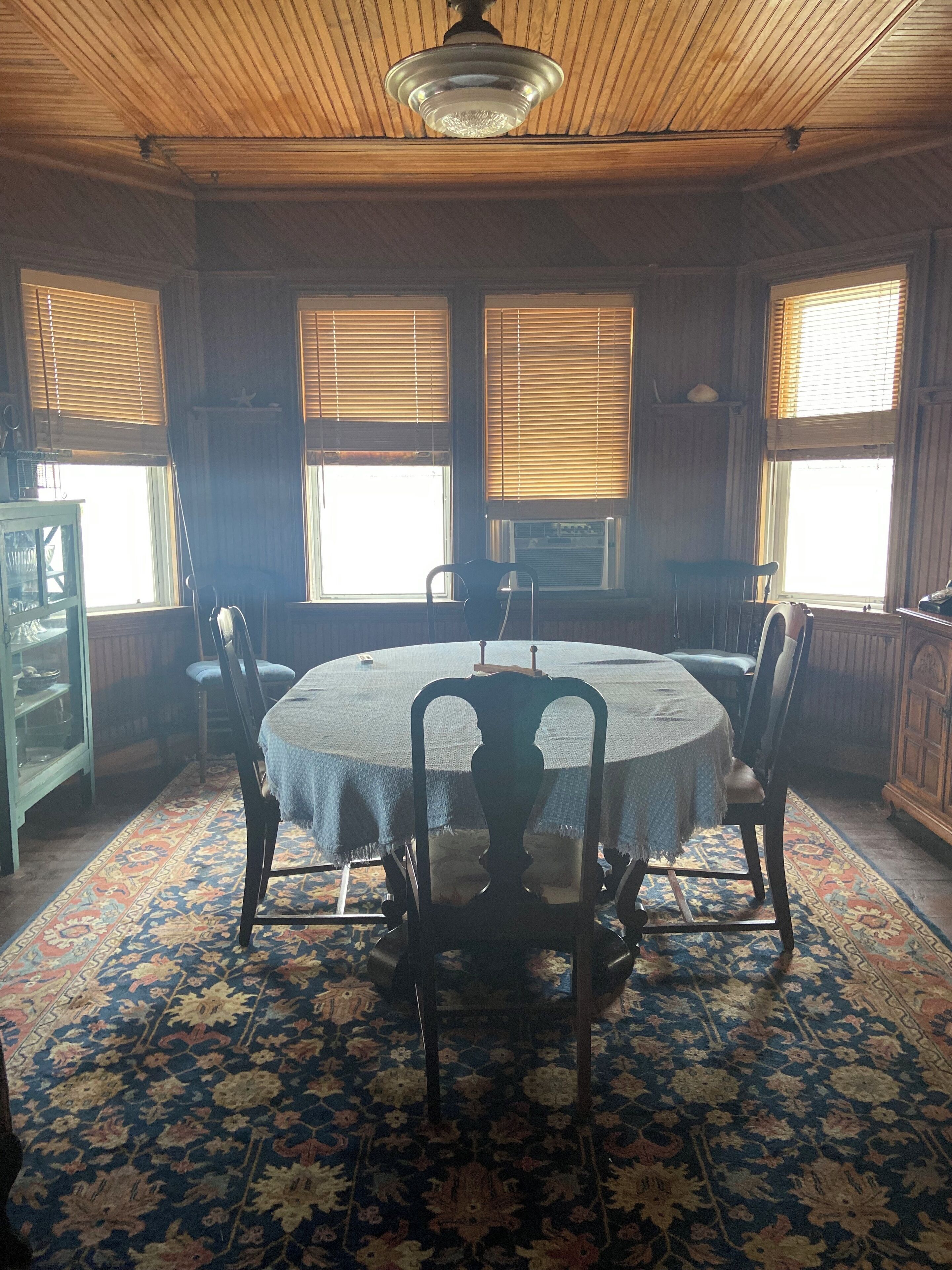 Une salle à manger avec une table ronde, quatre chaises et un tapis à motifs. On y trouve deux fenêtres équipées de stores, un ventilateur de plafond et un meuble avec une vitrine.