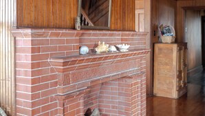 Ein Kamin aus Ziegelsteinen mit Dekorationen, ein Korbstuhl und ein Holzschrank in einem Raum mit Holzwänden und Holzdielenboden.
