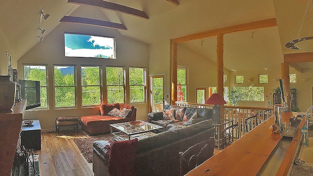 Un salón espacioso con una gran televisión montada en la pared, varias ventanas que ofrecen vistas a zonas verdes, un sofá rojo y una escalera de madera que conduce a un nivel superior.