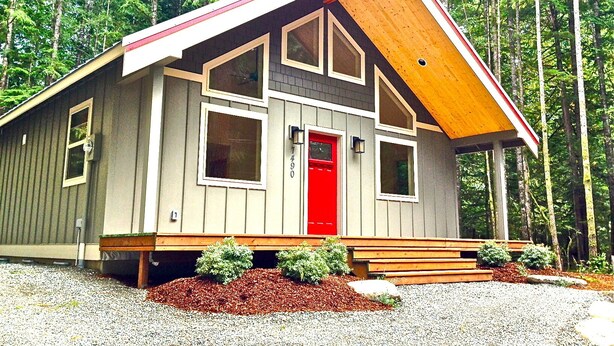 Una cabaña con puerta roja, ventanas amplias y techo inclinado, rodeada por un bosque.