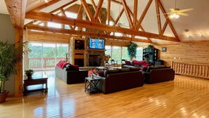A spacious living room with a high ceiling, wooden beams, a stone fireplace, and large windows offering a view of the outdoors. - Welcome to “Majestic View” – The View of a Lifetime! (Burnside)