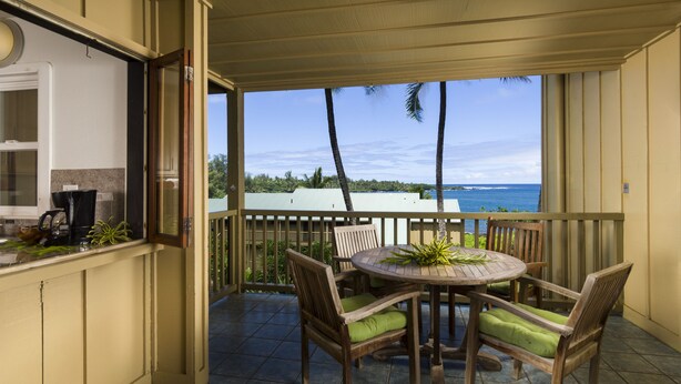 A balcony with a round wooden table and chairs, a view of the ocean, and a kitchen area with a coffee maker.