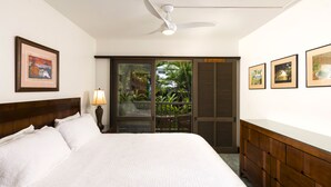 A hotel room with a bed, a ceiling fan, a nightstand with a lamp, a wooden dresser, and a sliding glass door leading to a balcony with plants.