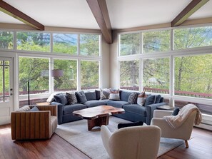 Una sala de estar moderna con un sofá seccional, una mesa de centro y un sillón, ventanas amplias con vista a árboles y vigas de madera en el techo.