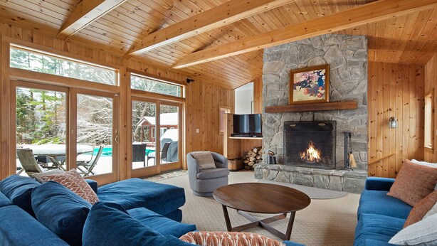 Un salón acogedor con chimenea de piedra, vigas de madera en el techo y ventanas amplias con vistas a un paisaje nevado.