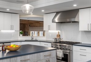 Una cocina moderna con armarios blancos, una isla grande con un cuenco de fruta, una estufa de acero inoxidable y paneles de madera en el fondo.