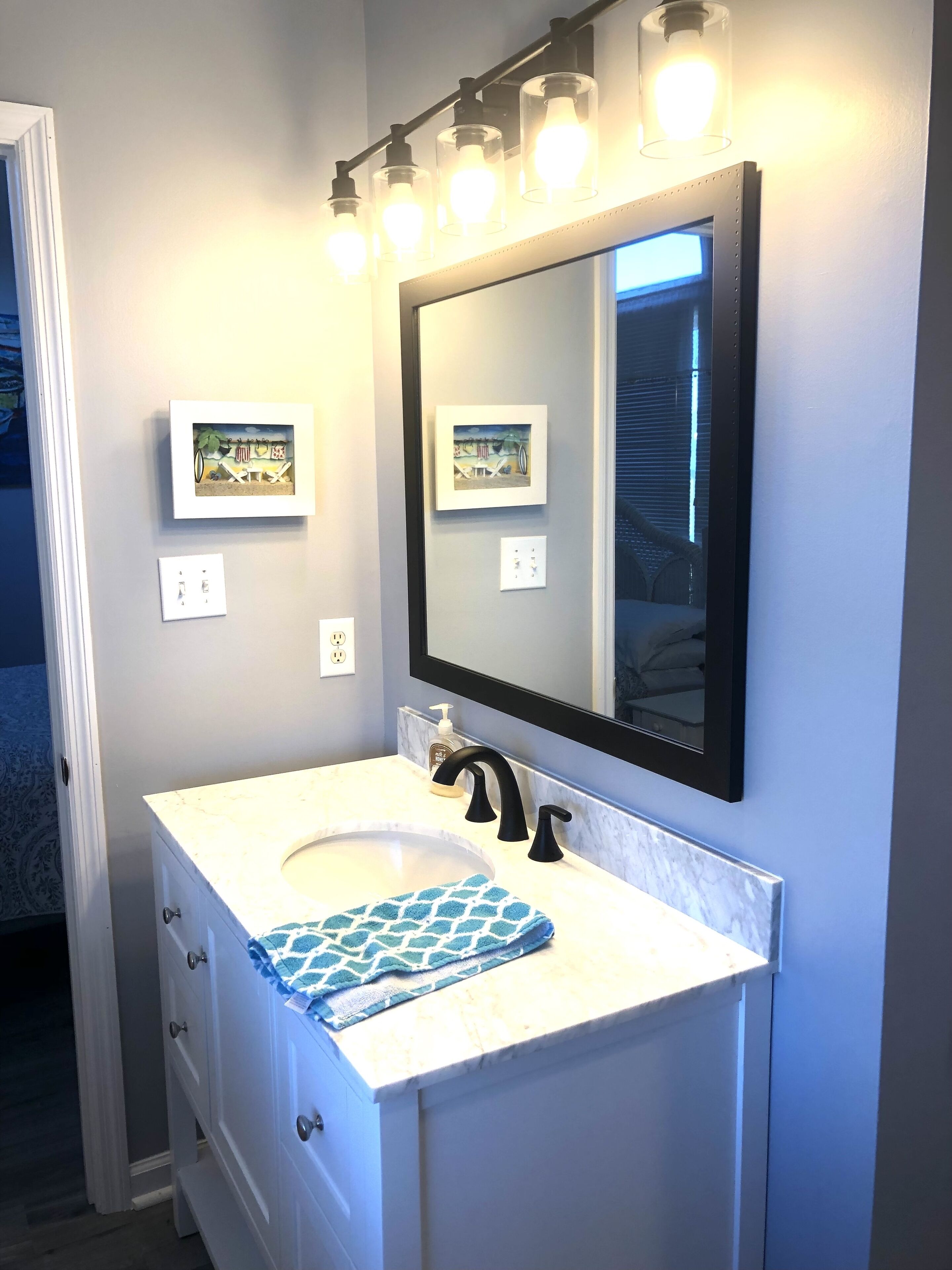 Une salle de bain avec un meuble-lavabo blanc, un robinet noir et un miroir surmonté de luminaires. Deux tableaux encadrés sont accrochés au mur.