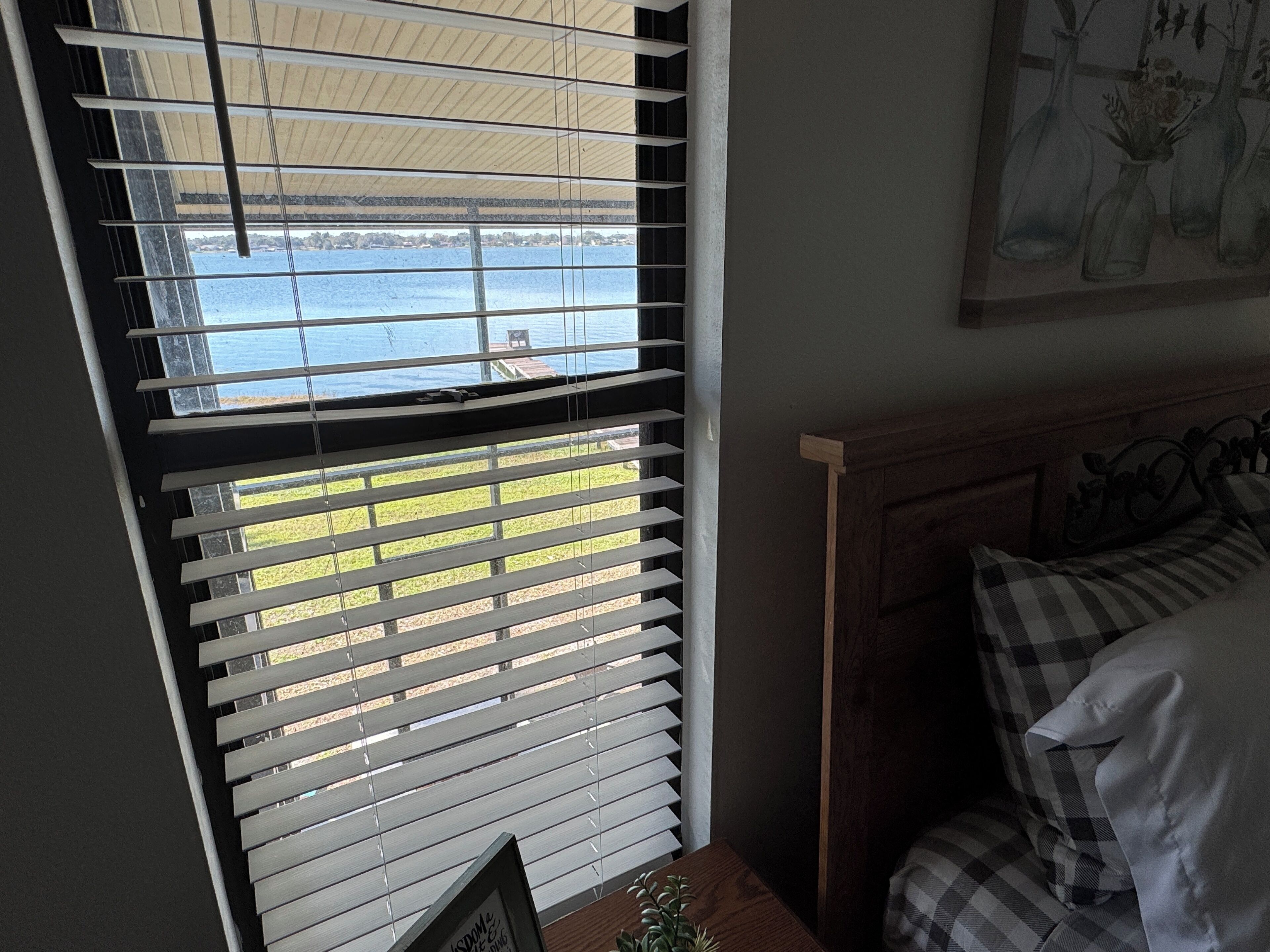 A room with a view of a body of water through partially open blinds, a bed with checkered bedding, and a framed picture on a small table.