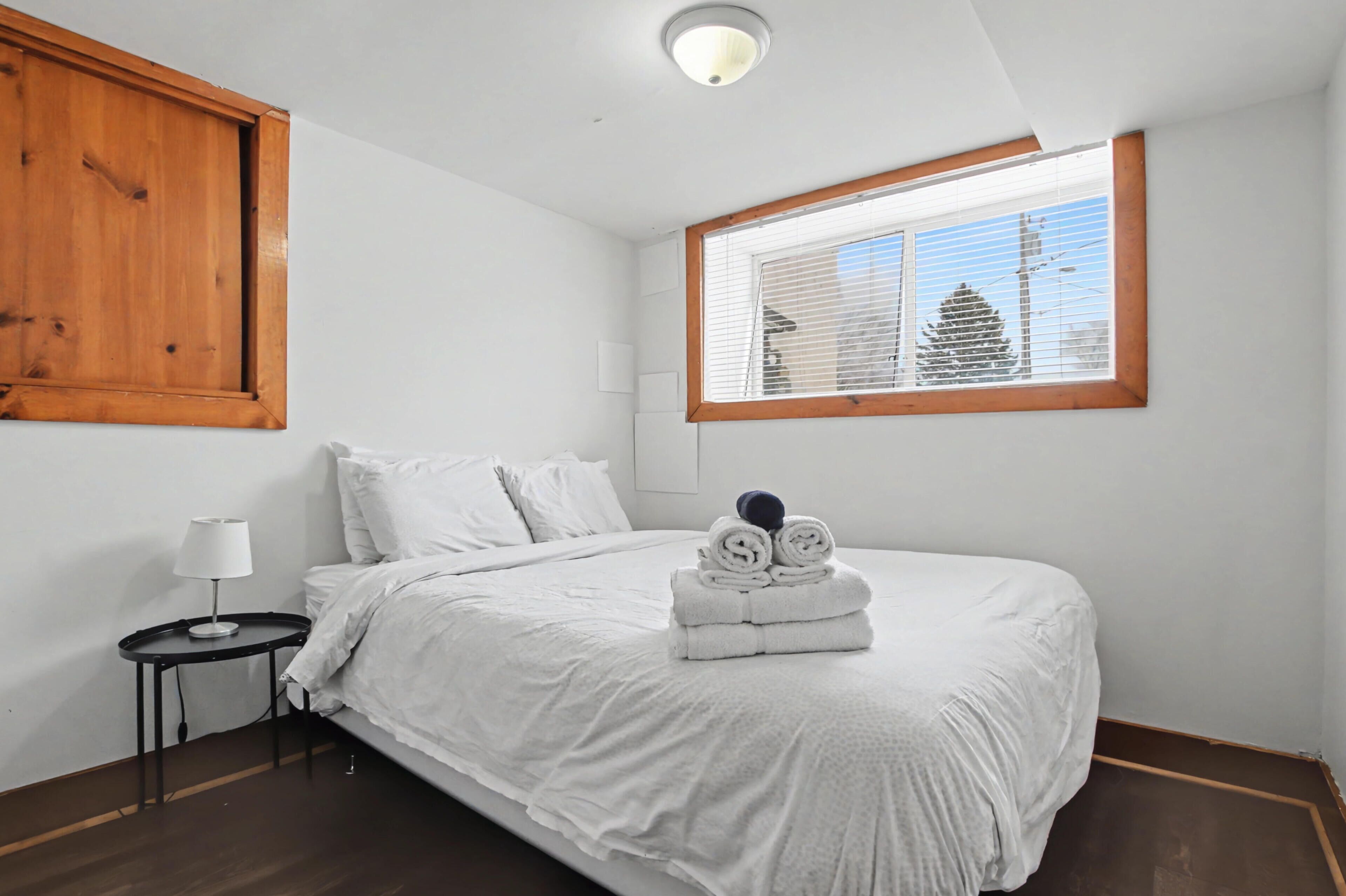 A neatly made bed with white linens, a small bedside table with a lamp, and a window with blinds.