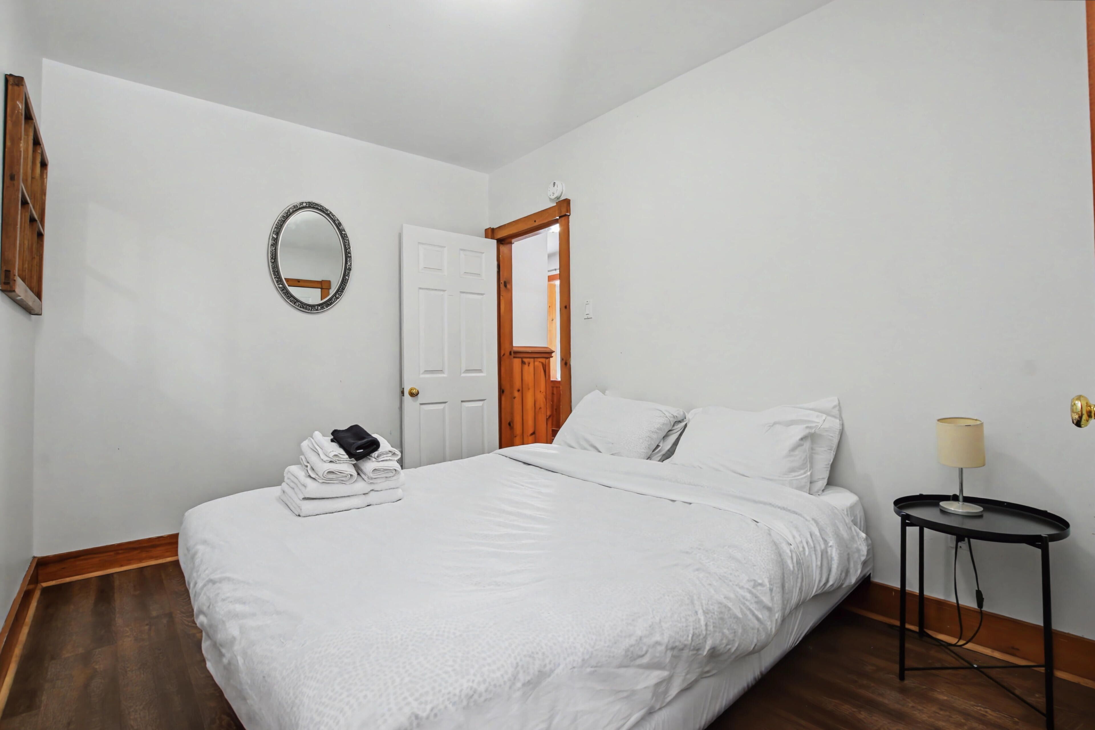 A bedroom with a bed, a bedside table with a lamp, a mirror, and a door leading to another room.
