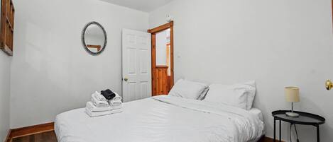A bedroom with a bed, a bedside table with a lamp, a mirror, and a door leading to another room.