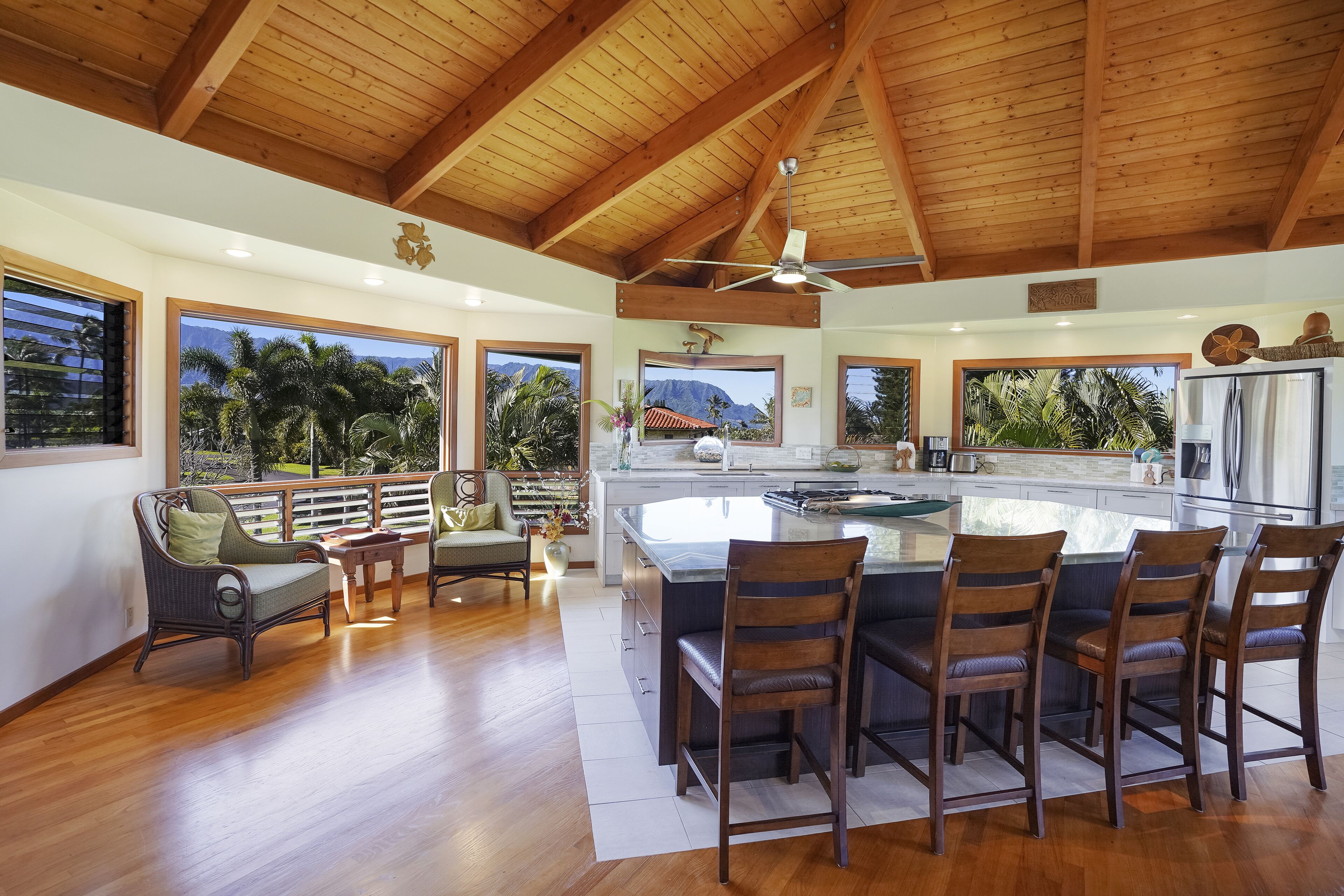 Una cocina amplia con una isla grande, vigas de madera en el techo y ventanas grandes que ofrecen vistas a palmeras y montañas.