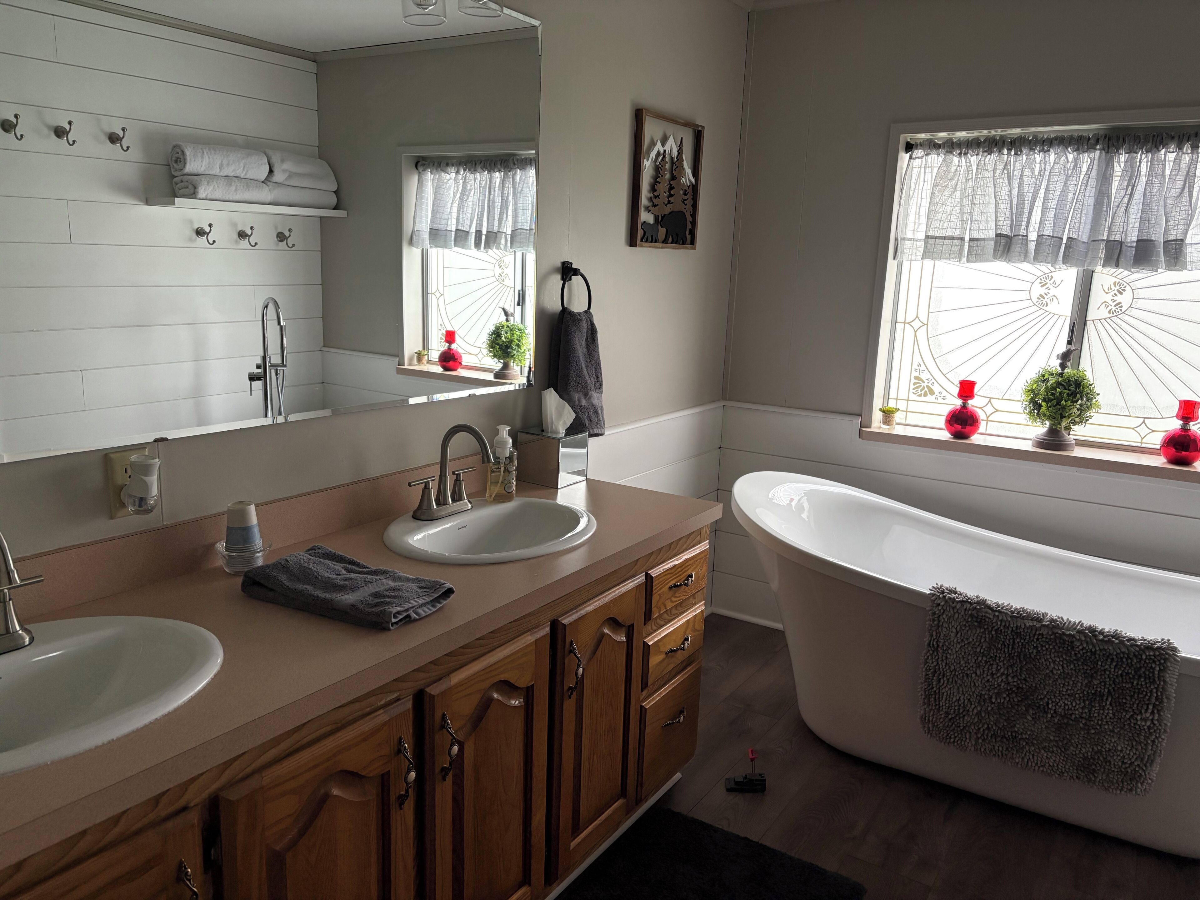A modern bathroom with two sinks, a bathtub, wooden cabinets, a mirror, and a window with decorative curtains.