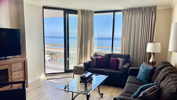 A modern living room with a glass coffee table, a sofa with patterned cushions, a TV on a wooden cabinet, and a view of the ocean through large windows. - Oceanfront Dream Vacation! 11th Floor-What a View!! + Beach Locker-Book Early! (Myrtle Beach)