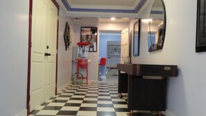 A hallway with a black and white checkered floor, a white door, a red vending machine, a mirror, and a framed picture on the wall.