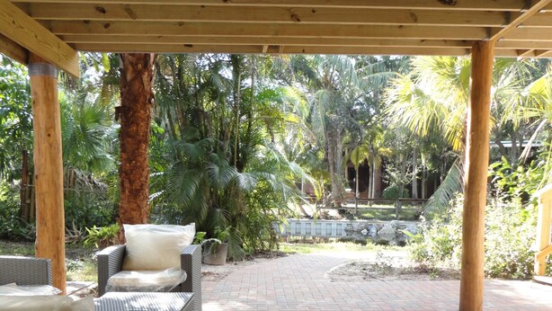 An outdoor patio with wicker furniture and a brick floor, surrounded by tropical vegetation.
