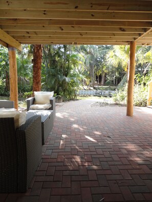 Un patio al aire libre con mobiliario de mimbre y piso de ladrillo, rodeado de follaje tropical.