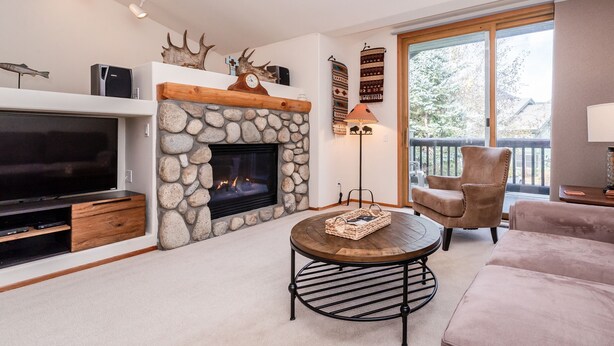 A living room with a stone fireplace, a television, a round coffee table, and a large window with a view of trees. - Home Away From Home in Snowcreek V Resort (Mammoth Lakes)