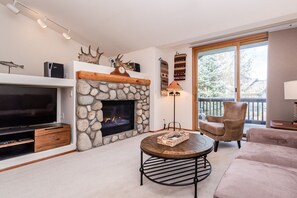 A living room with a stone fireplace, a television, a round coffee table, and a large window with a view of trees. - Home Away From Home in Snowcreek V Resort (Mammoth Lakes)