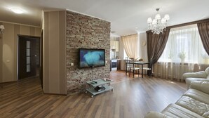 A living room with a flat-screen TV mounted on a brick wall, a leather sofa, a dining area with a table and chairs, and a chandelier. - Clean, Safe and Affordable (Pechers'kyi district)
