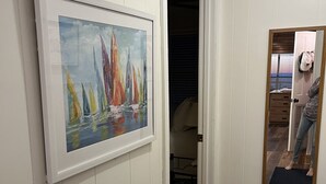 A hallway with a framed painting of sailboats, wooden flooring, and a mirror reflecting a person. - Sandy Beach, gorgeous sunsets, great front porch view! (Fairhope)