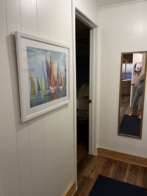 A hallway with a framed painting of sailboats, wooden flooring, and a mirror reflecting a person.