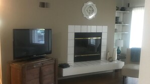 A living room with a flat-screen TV on a wooden cabinet, a white fireplace with a decorative wall piece above it, and a bookshelf with various items.