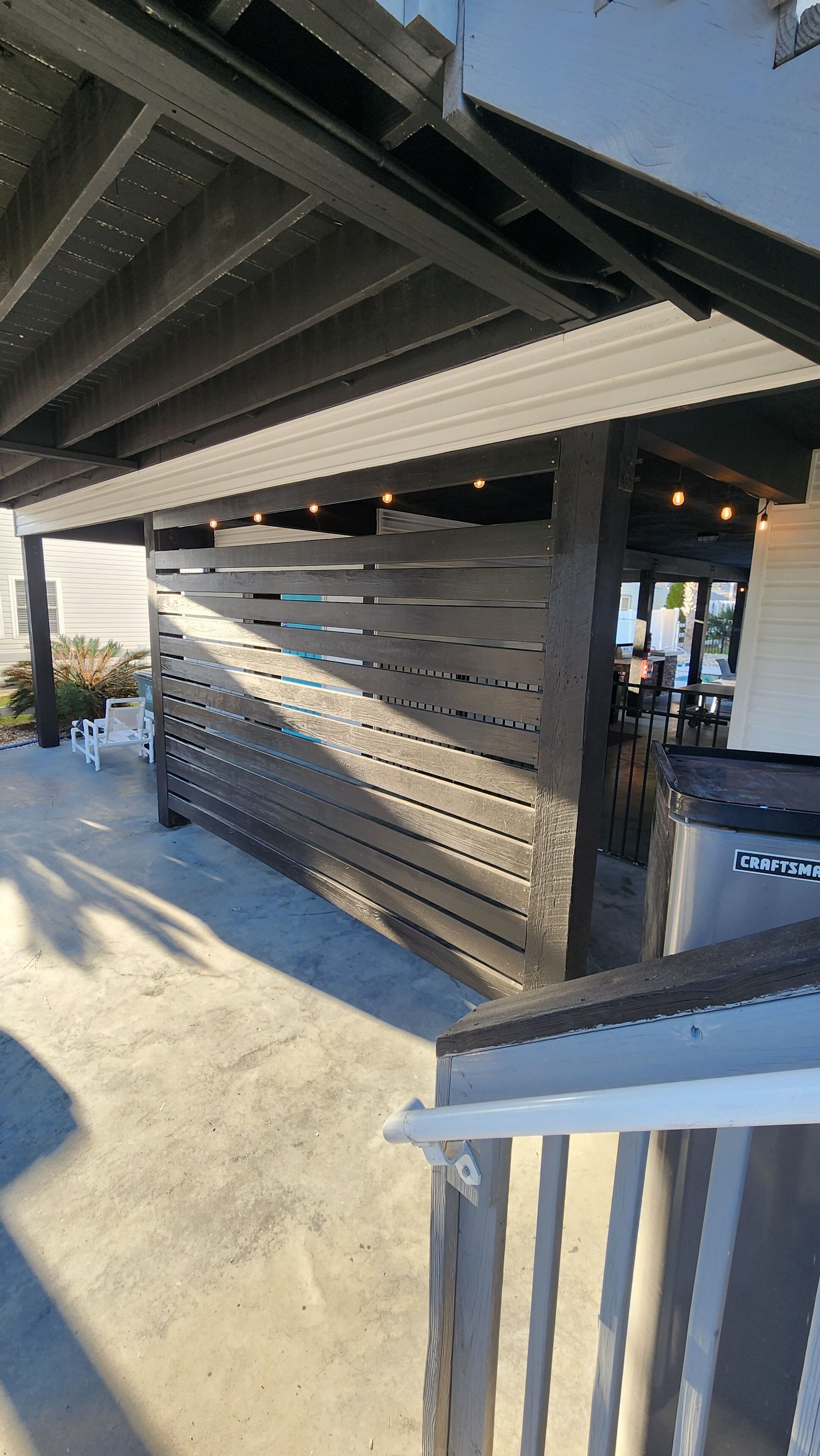 Un espacio al aire libre techado con un muro de listones de madera, suelo de hormigón y baranda de metal.
