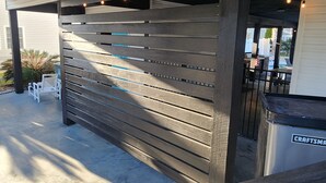 Ett takförsett utomhusområde med en vägg av träribbor, ett betonggolv och ett metallräcke.