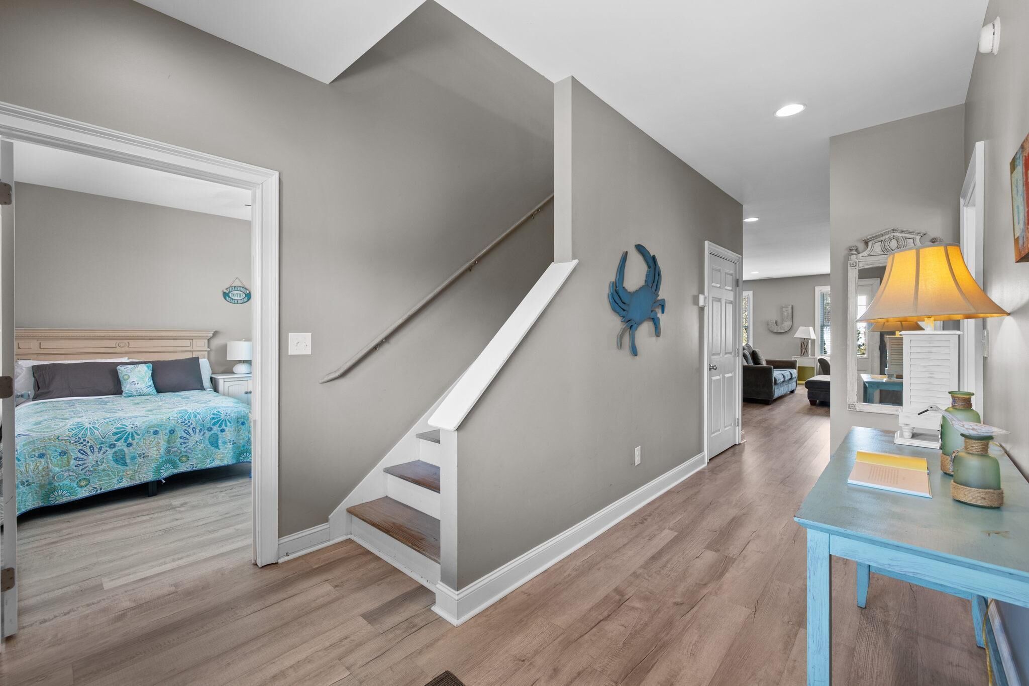 A modern interior with a staircase, a bed with patterned bedding, a blue wall decoration, and a table with a lamp.