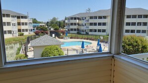 A view of a swimming pool surrounded by a fence, with a small building in the foreground and multi-story buildings in the background. - 1206 Palms, 3-bedroom, 2-bath condo, Sept dates at last minute & off-season rate (Rehoboth Beach)