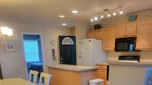 A kitchen with wooden cabinets, a refrigerator, a microwave, and a dining area with a table and chairs. - 1206 Palms, 3-bedroom, 2-bath condo, Sept dates at last minute & off-season rate (Rehoboth Beach)