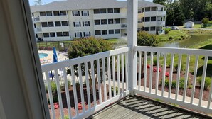 A wooden deck with a white railing, overlooking a landscaped garden with red flowers and a swimming pool. - 1206 Palms, 3-bedroom, 2-bath condo, Sept dates at last minute & off-season rate (Rehoboth Beach)