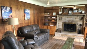 A cozy living room with a fireplace, leather sofas, wooden walls, and a bookshelf.